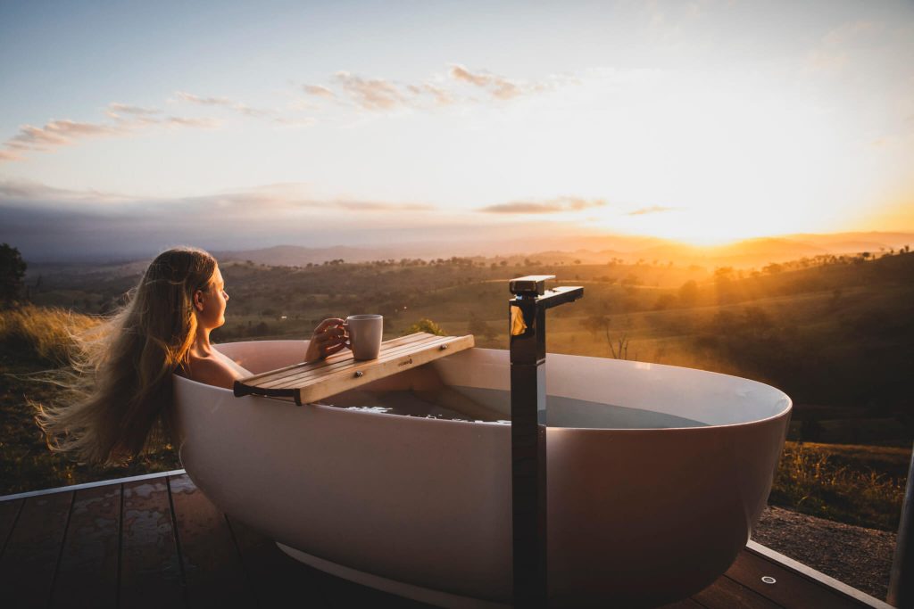 Beautiful lady with flowing blond hair laying back and soaking in a white outdoor bathtub enjoying a golden sunrise with coffee cup in hand.